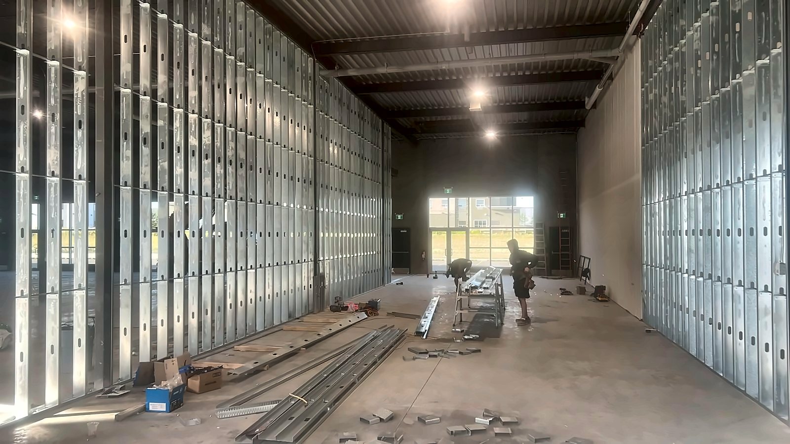 Two tradespeople cutting steel studs inside a long bay framed with galvanized steel stud walls