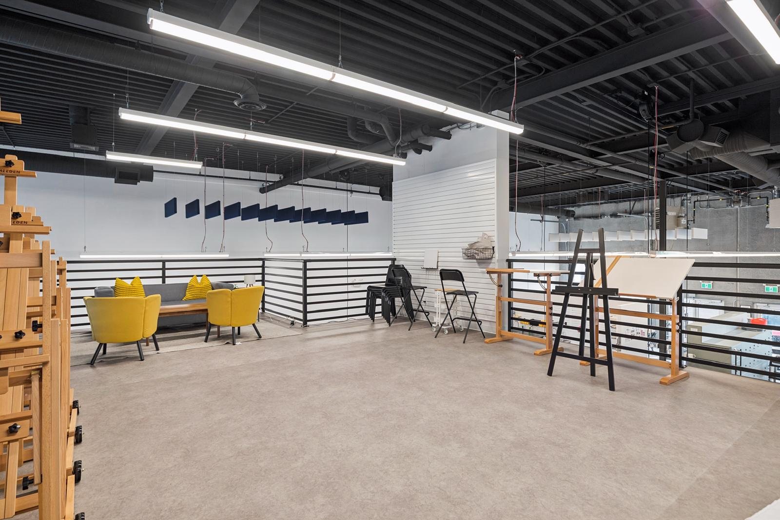 The mezzanine: yellow lounge chairs, drafting tables and easels behind a black steel railing under exposed ceiling